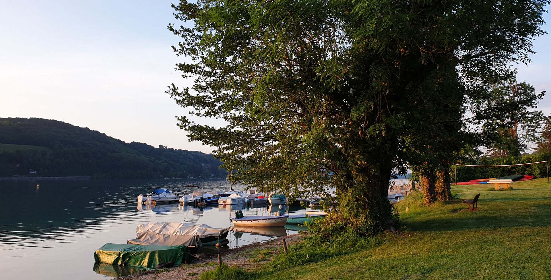 Vue depuis la plage du bord du lac. La pelouse de la plage, le banc, pour regarder le lac de Paladru, la table de ping-pong, le terrain de volley. Les bateaux amarrés sur l'eau.